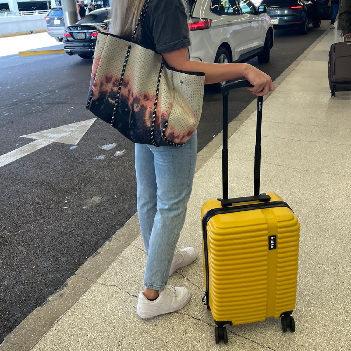 Person with a yellow suitcase and patterned tote bag on a sidewalk.