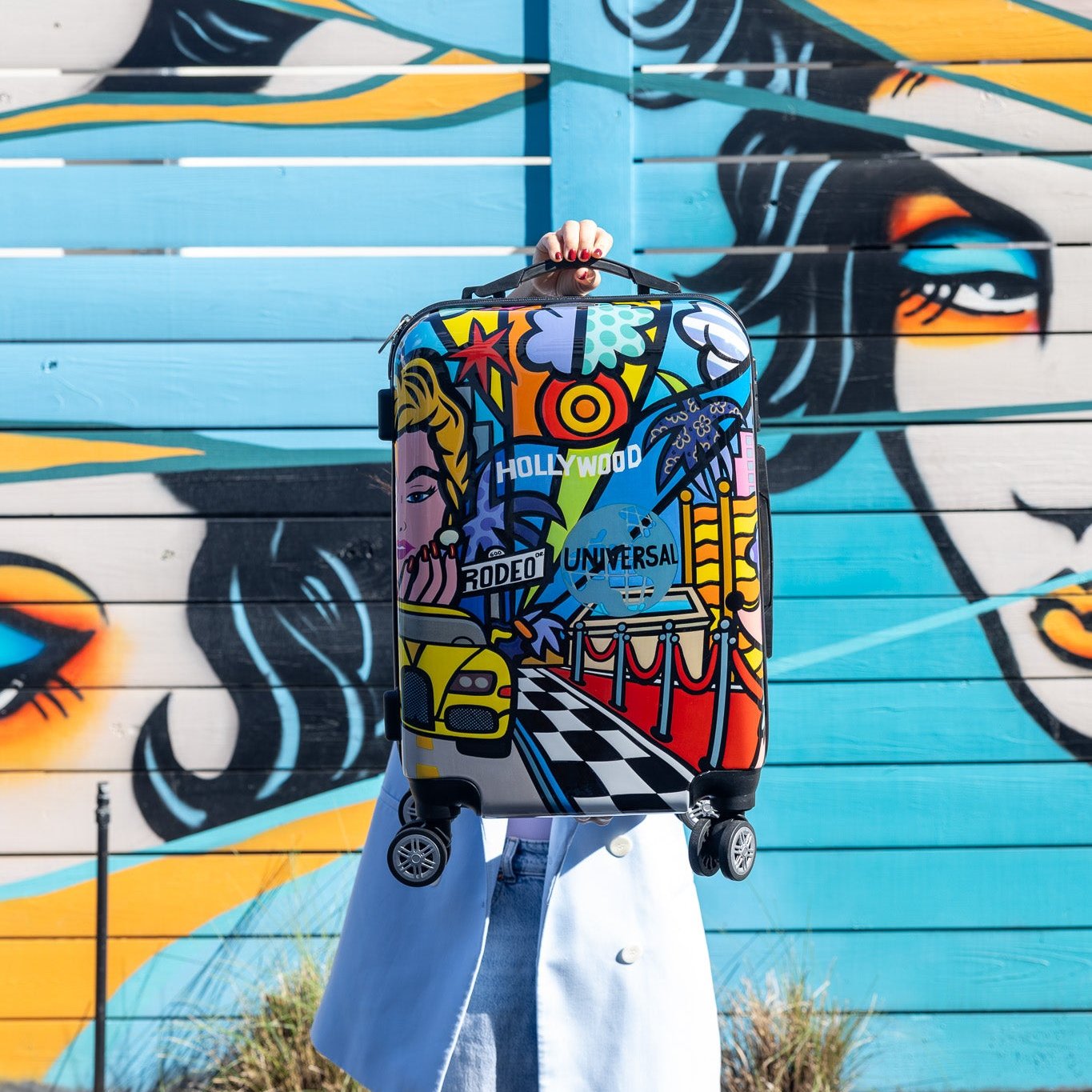 Person holding a colorful suitcase with a Hollywood-themed design against a graffiti-covered wall.