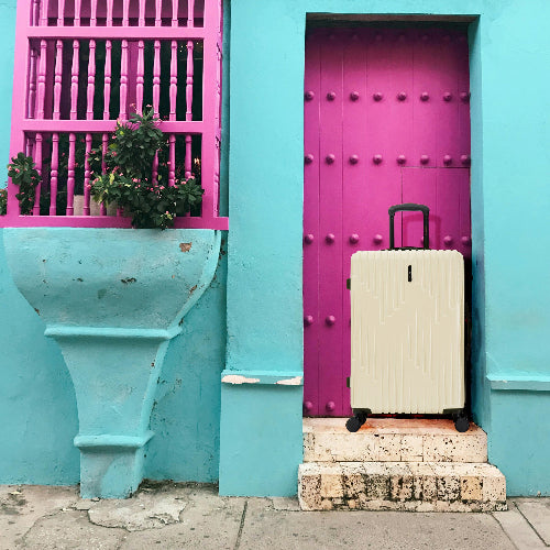 Beige suitcase on a stone step in front of a turquoise wall with a pink door and balcony.