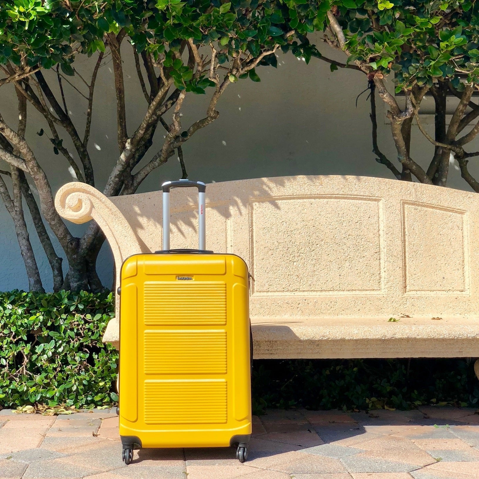 Yellow suitcase on a stone path with a bench and trees in the background