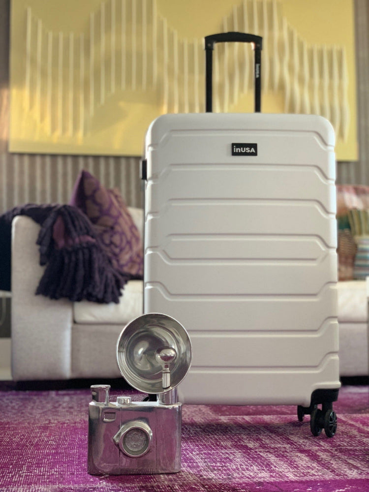 White suitcase with 'inUSA' branding on a purple rug, with a blurred indoor background.