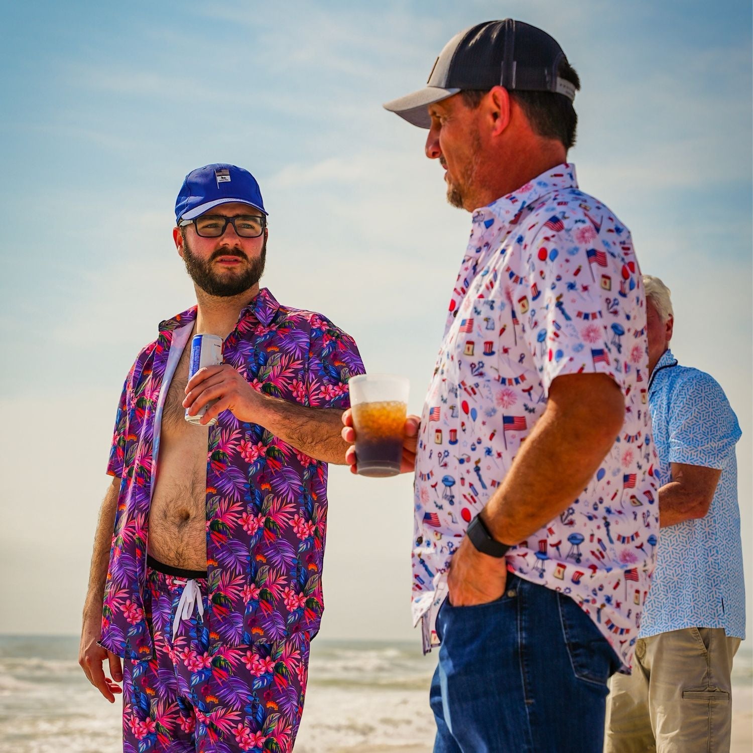 Two men on a beach wearing colorful shirts and hats, holding drinks.