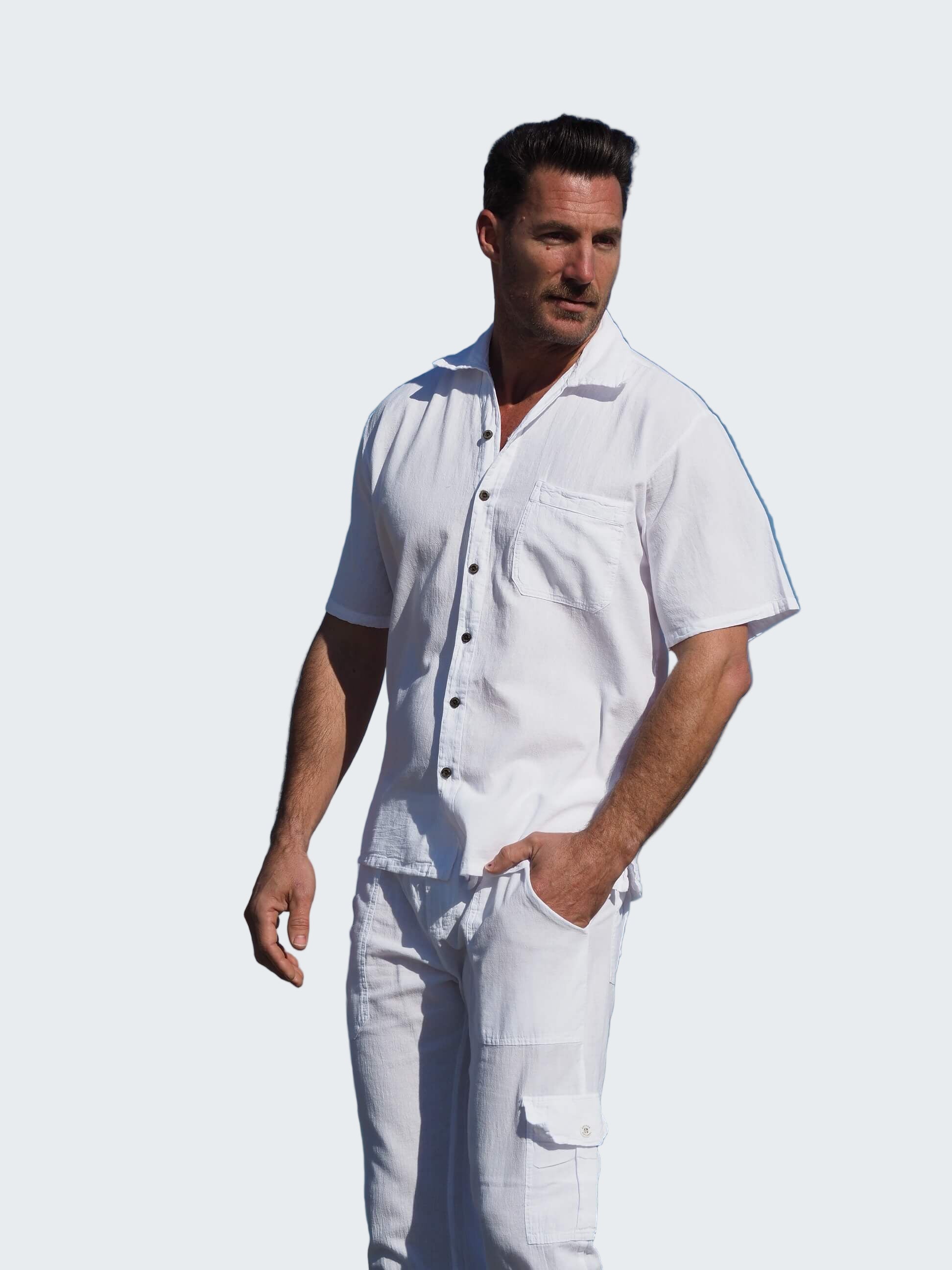 Man in a white outfit standing on a beach with palm trees and blue sky.