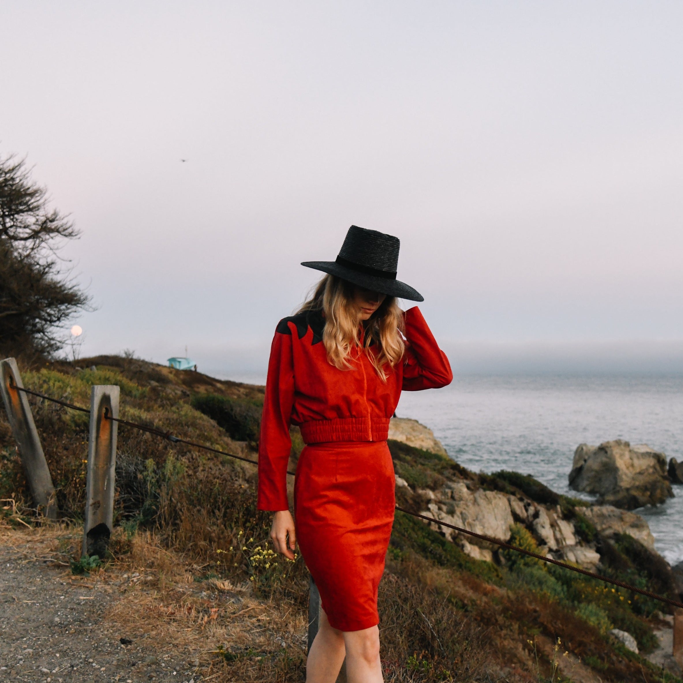 Woman in a red outfit and black hat standing on a coastal path with ocean view.