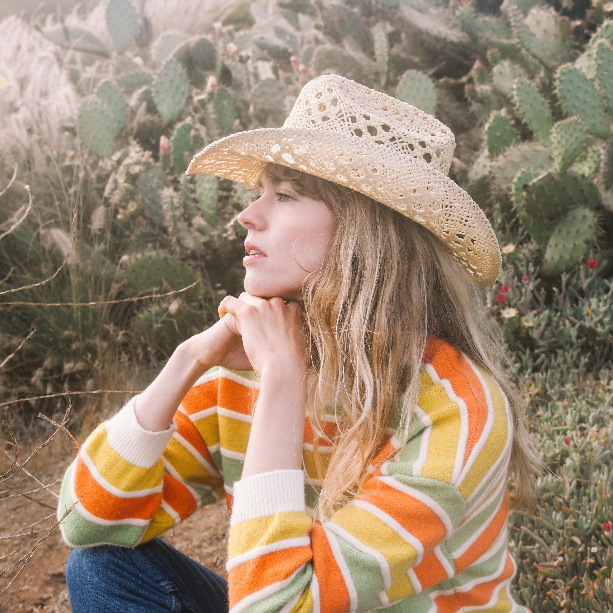 Woman wearing a straw hat and colorful sweater in a desert setting with cacti.