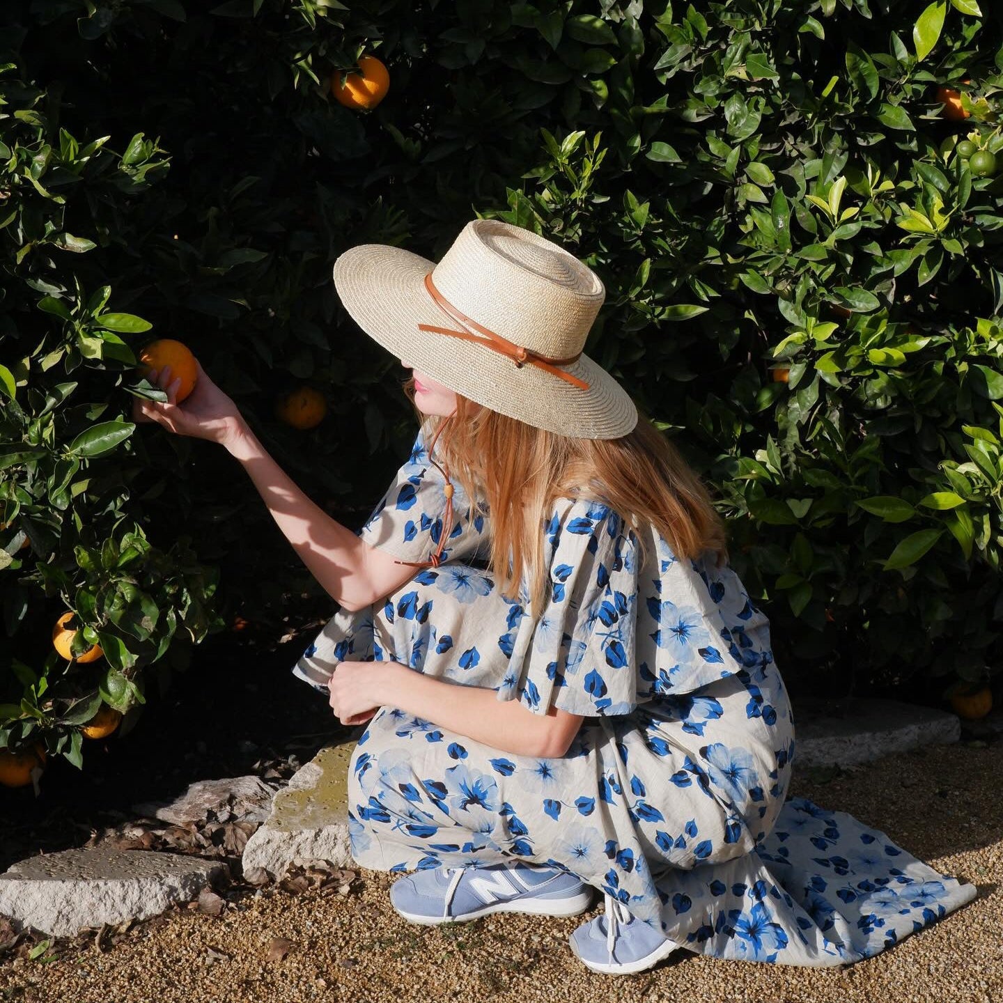 Woman in a floral dress and sun hat picking oranges from an orange tree.