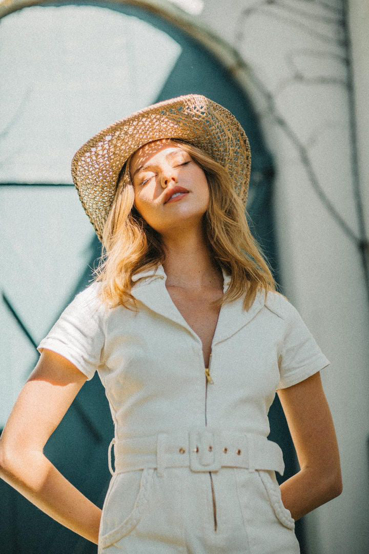Woman wearing a straw hat and white outfit against a textured wall.