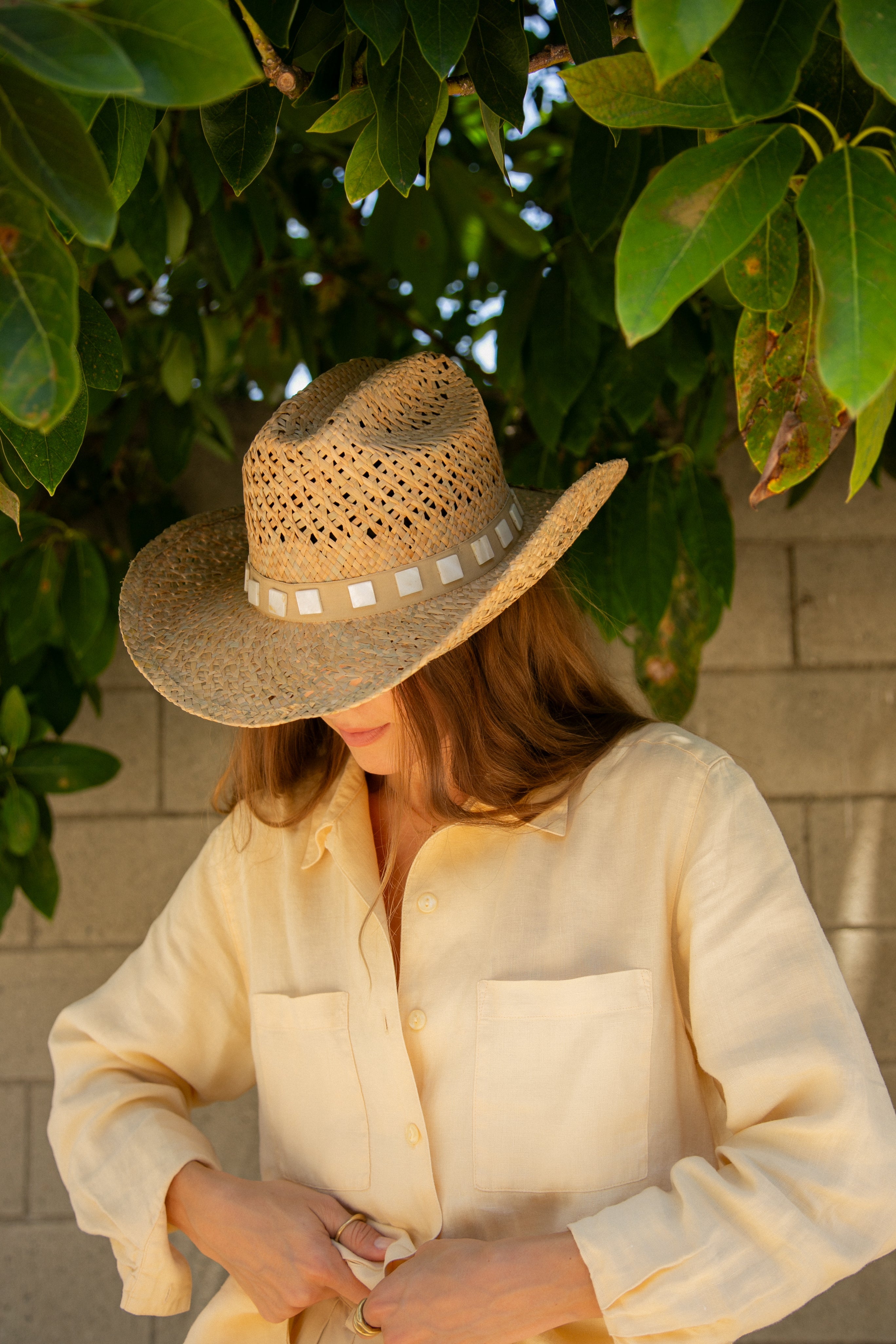 Person wearing a straw hat and light-colored shirt sitting under green leaves.