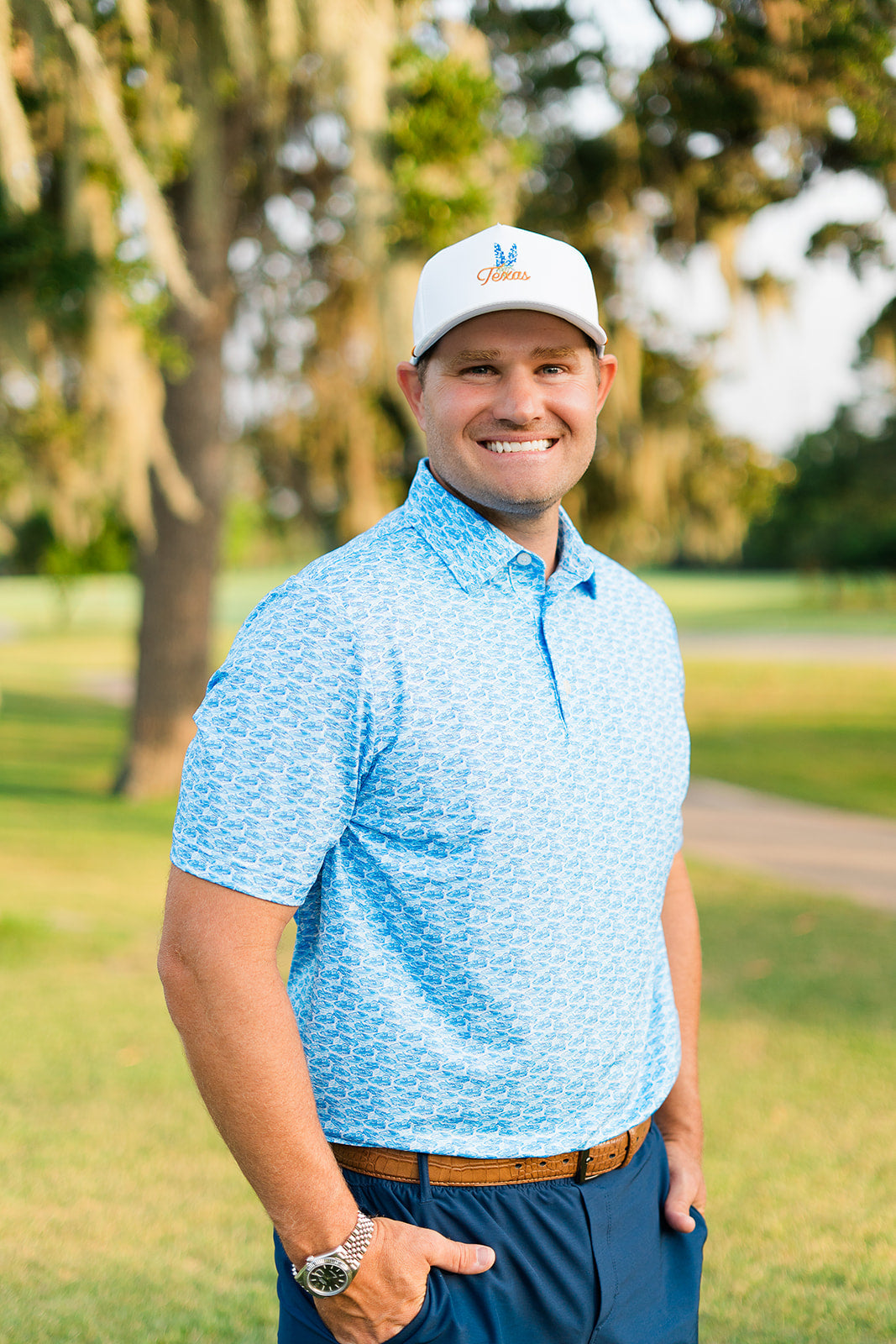 Man wearing a blue shirt and white cap standing on a grassy area with trees in the background