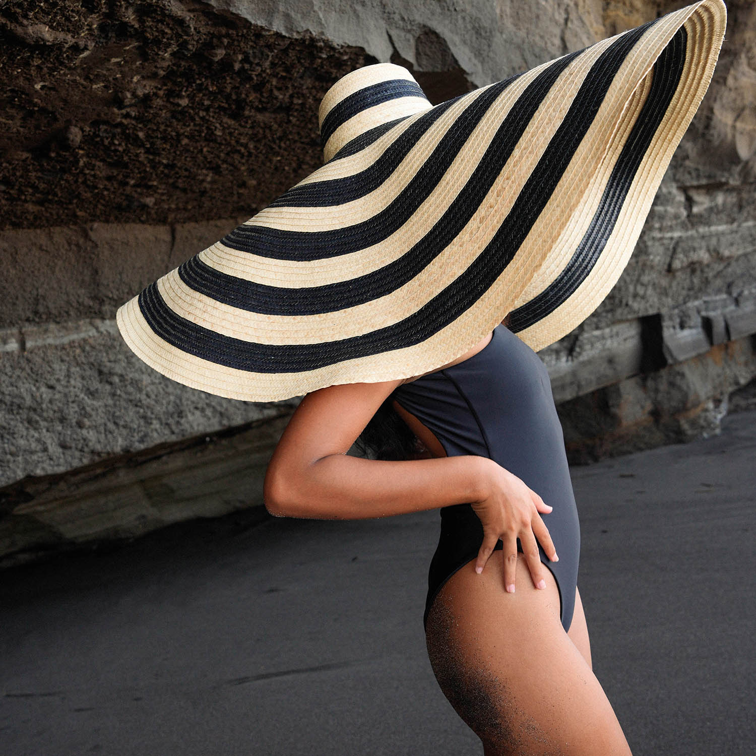 Person wearing a large black and beige striped sun hat against a stone wall.