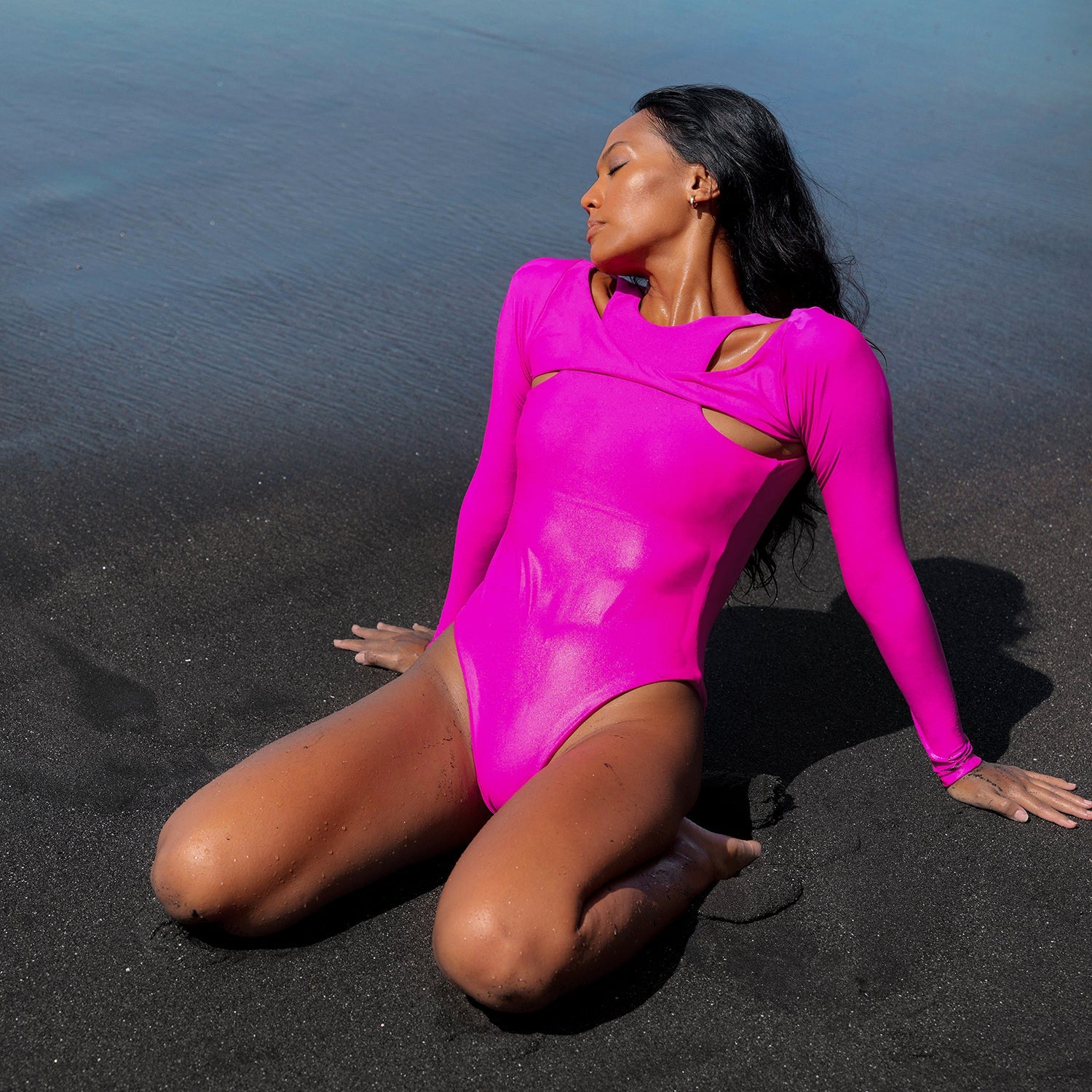 Woman in a bright pink bodysuit sitting on black sand with water in the background