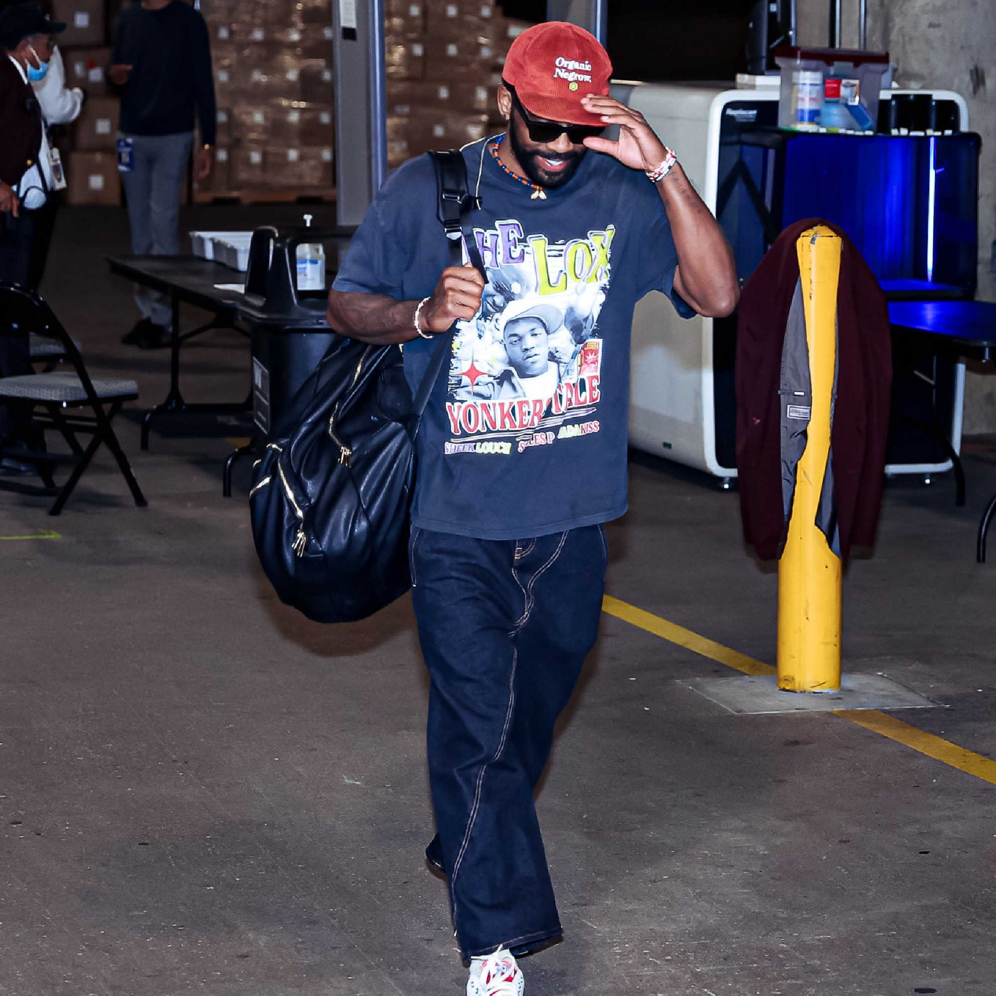 Man wearing a graphic t-shirt and red cap walking in an indoor setting with tables and chairs.