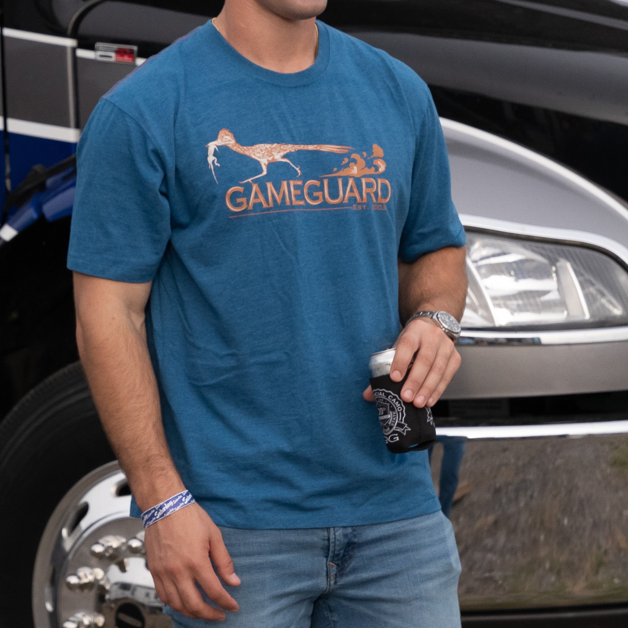 Person wearing a blue 'Gameguard' t-shirt holding a coffee cup in front of a vehicle.