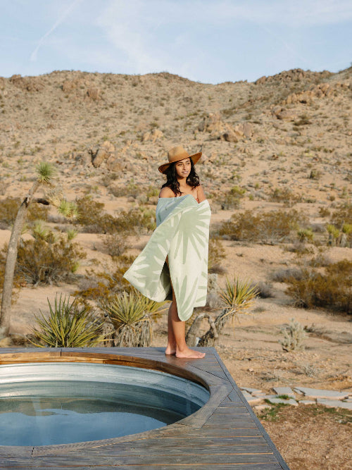 Woman wrapped in a light green towel standing by a small pool in a desert landscape.