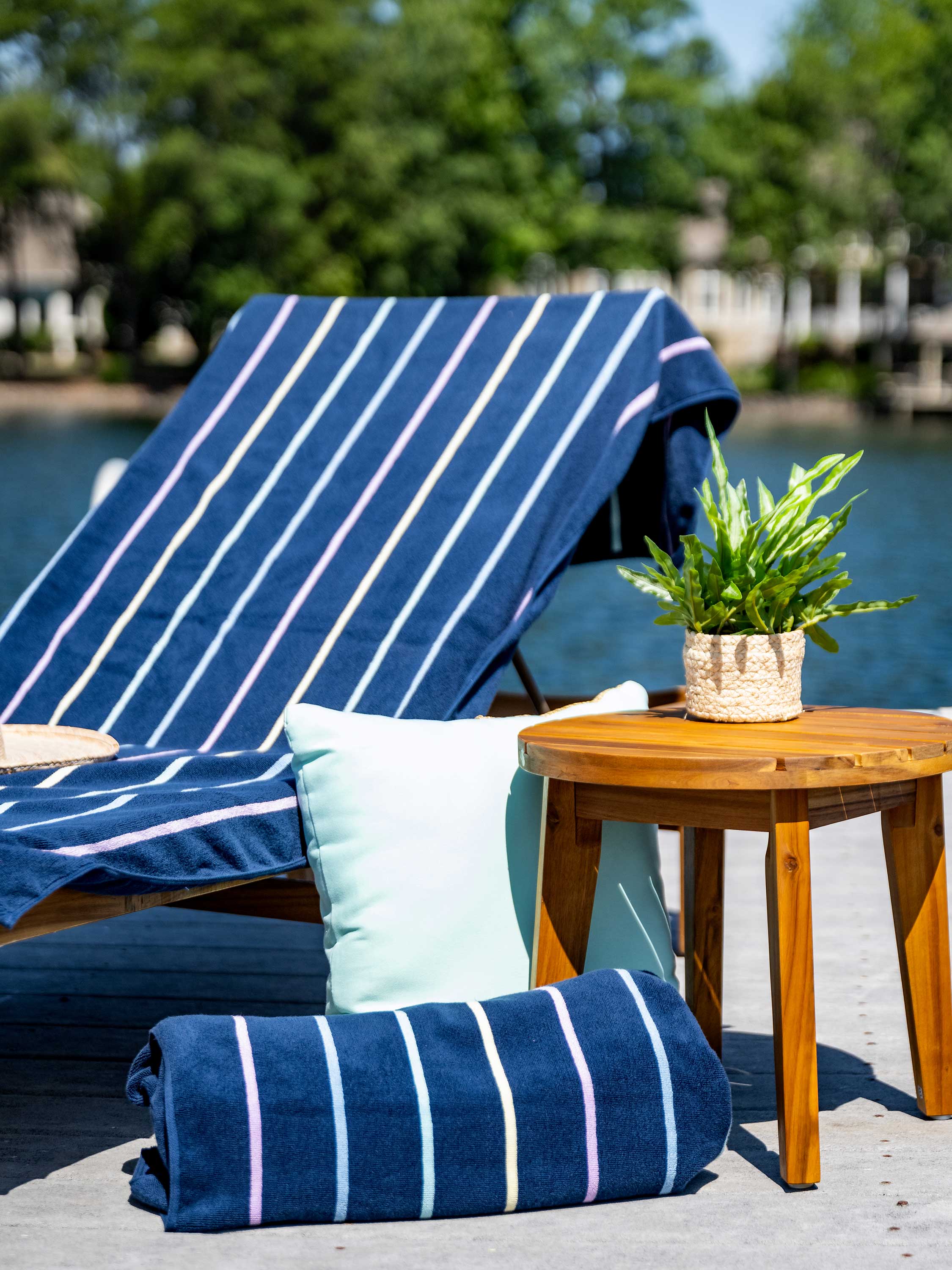 Navy striped towel on a chair with a wooden table and plant by a waterfront.