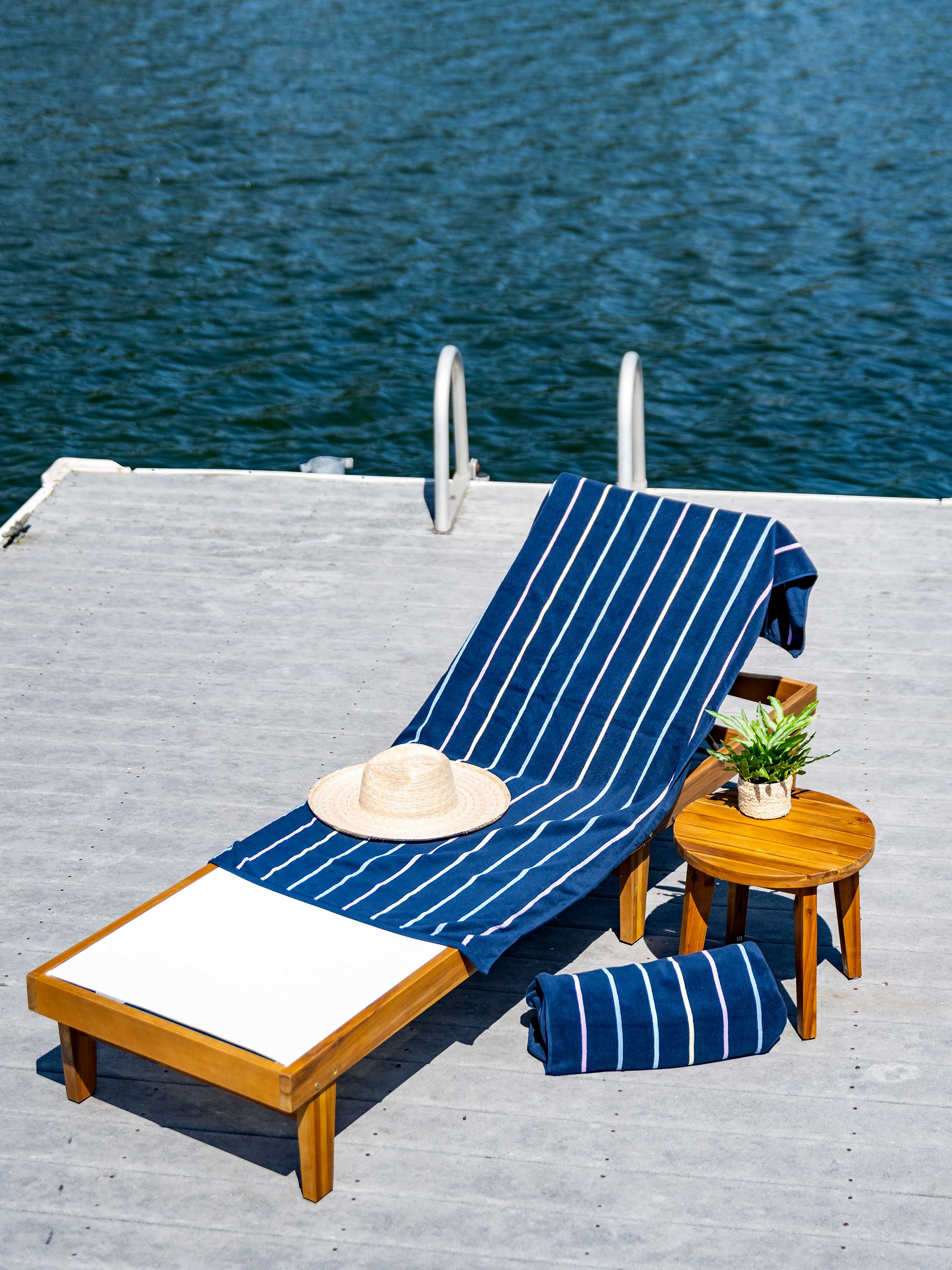 Wooden lounge chair with blue striped cover and white cushion on a dock by water.