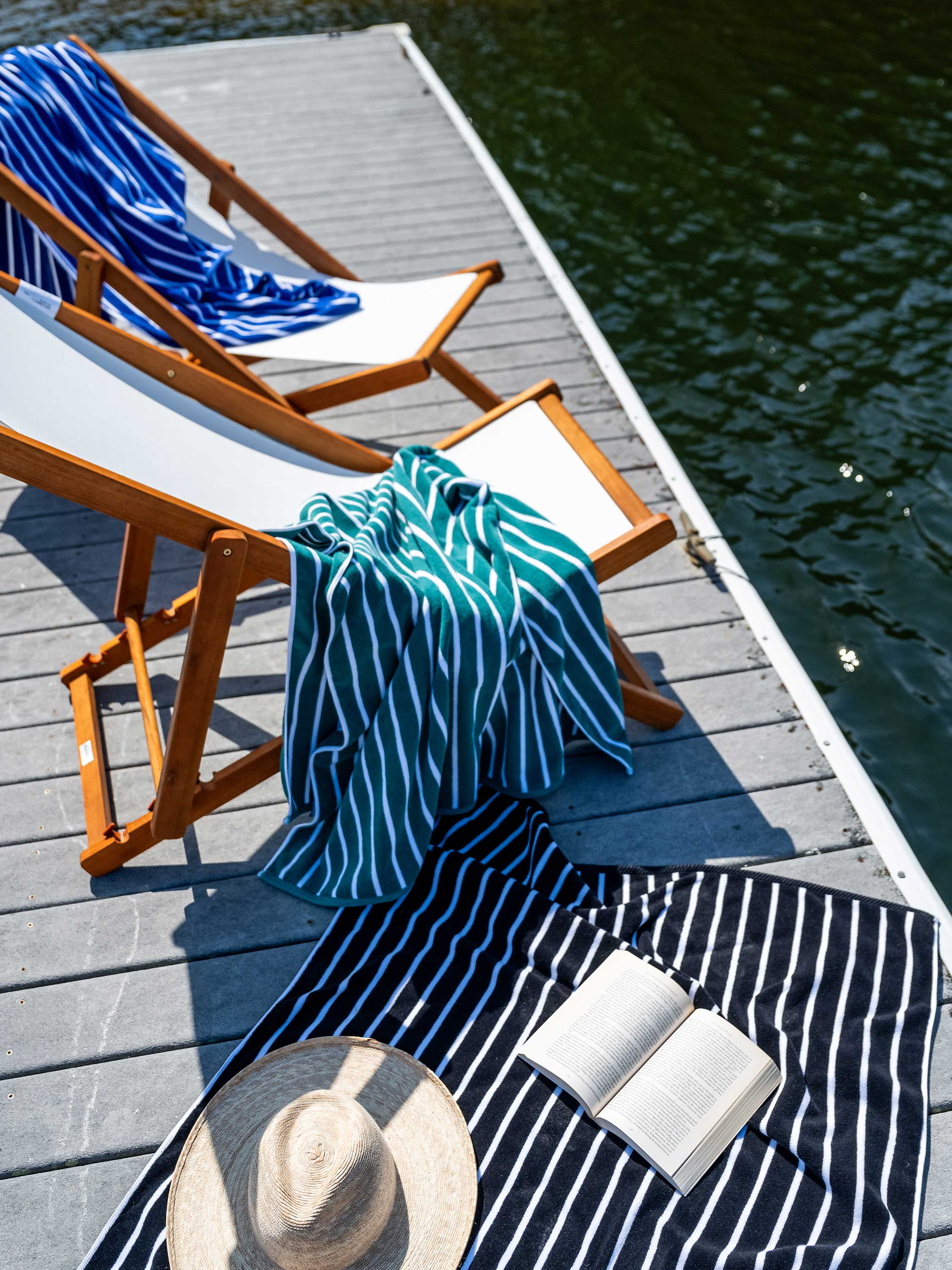 Two wooden lounge chairs with striped towels and a hat on a dock by water.