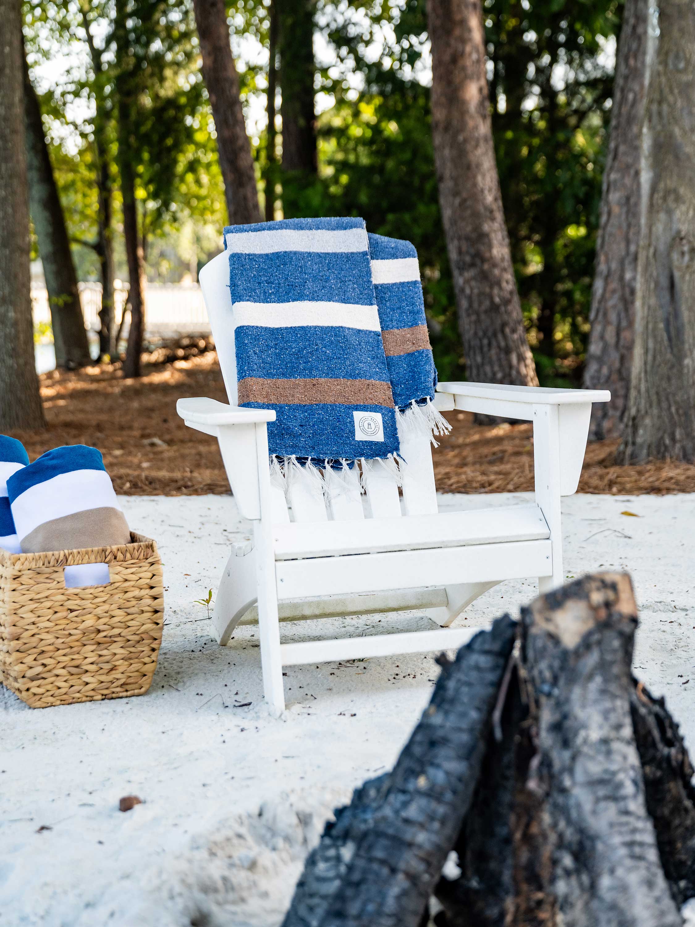 White Adirondack chair with a blue and white striped towel on a sandy beach with trees in the background.