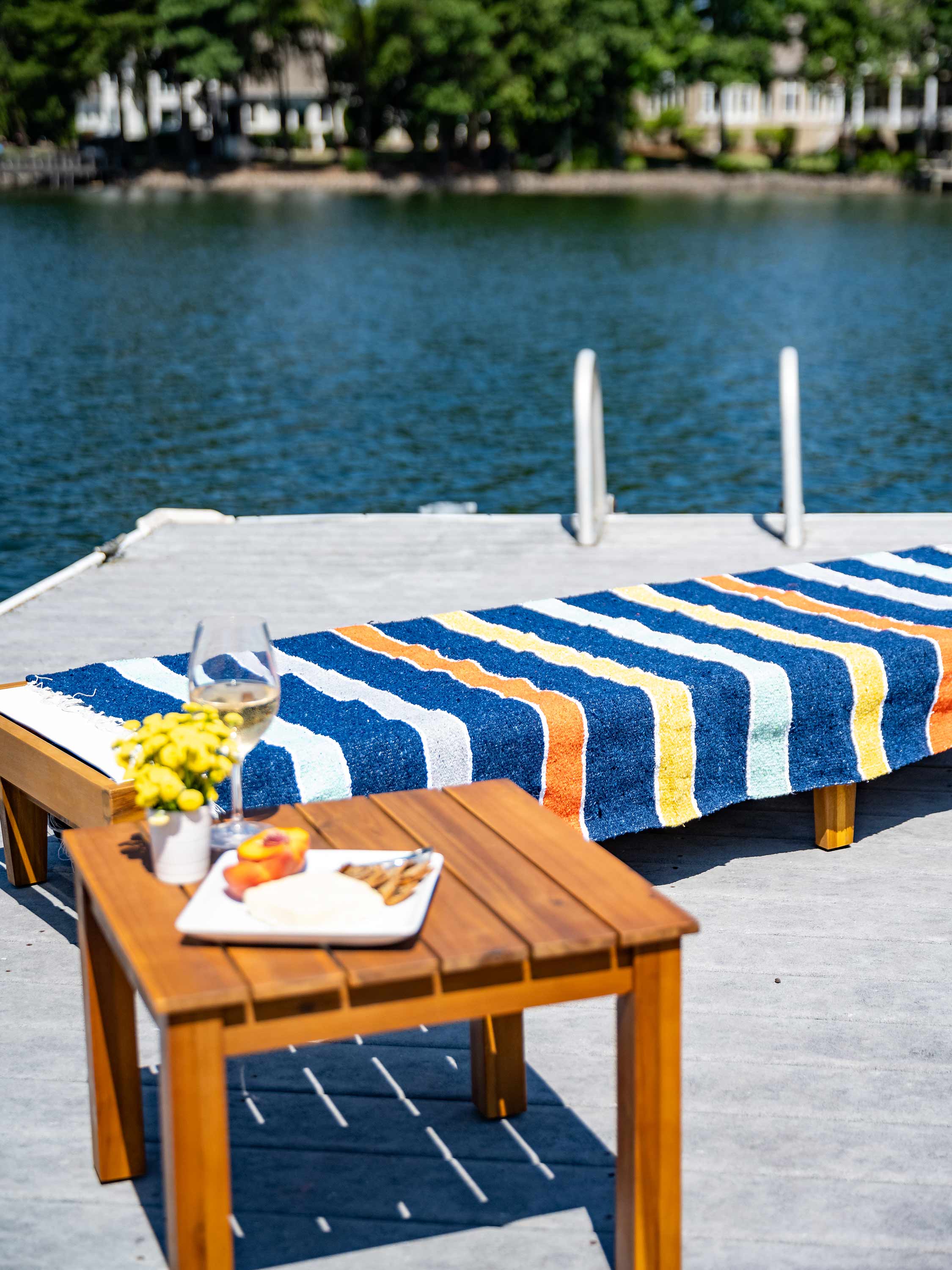 Outdoor setting with a striped towel on a chair, wooden table with food and drinks, and water in the background.
