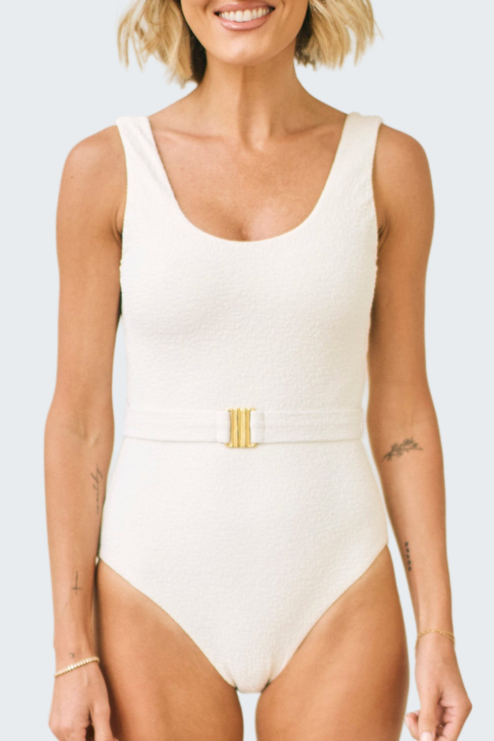 Woman wearing a white one-piece swimsuit with a gold belt on a plain background