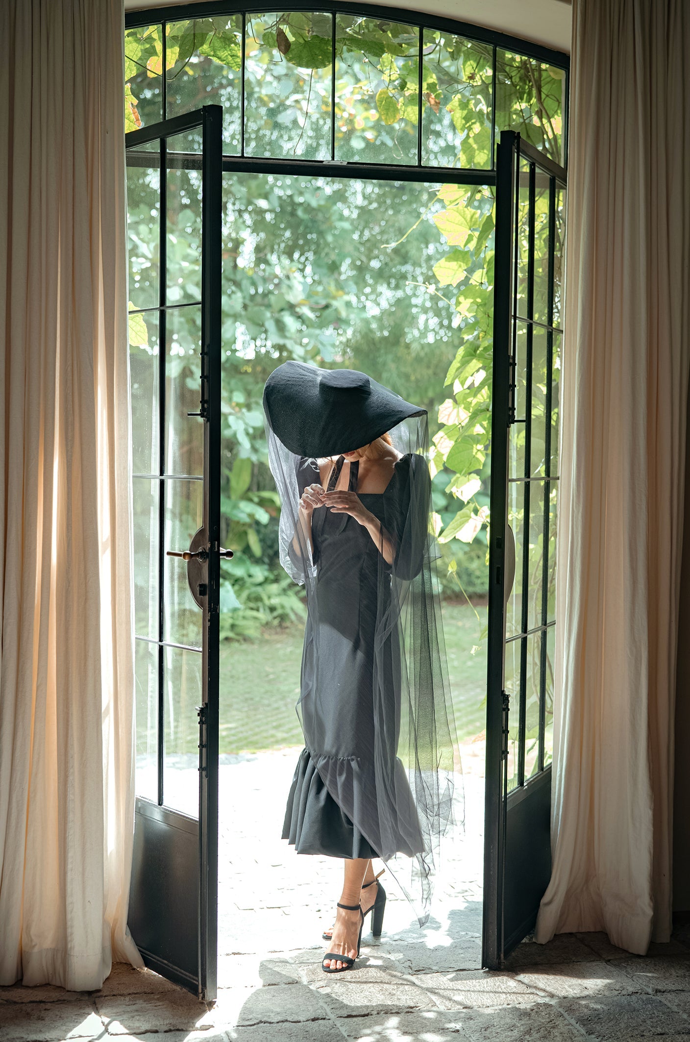 Woman in a black dress and hat standing in a doorway with white curtains and greenery outside.
