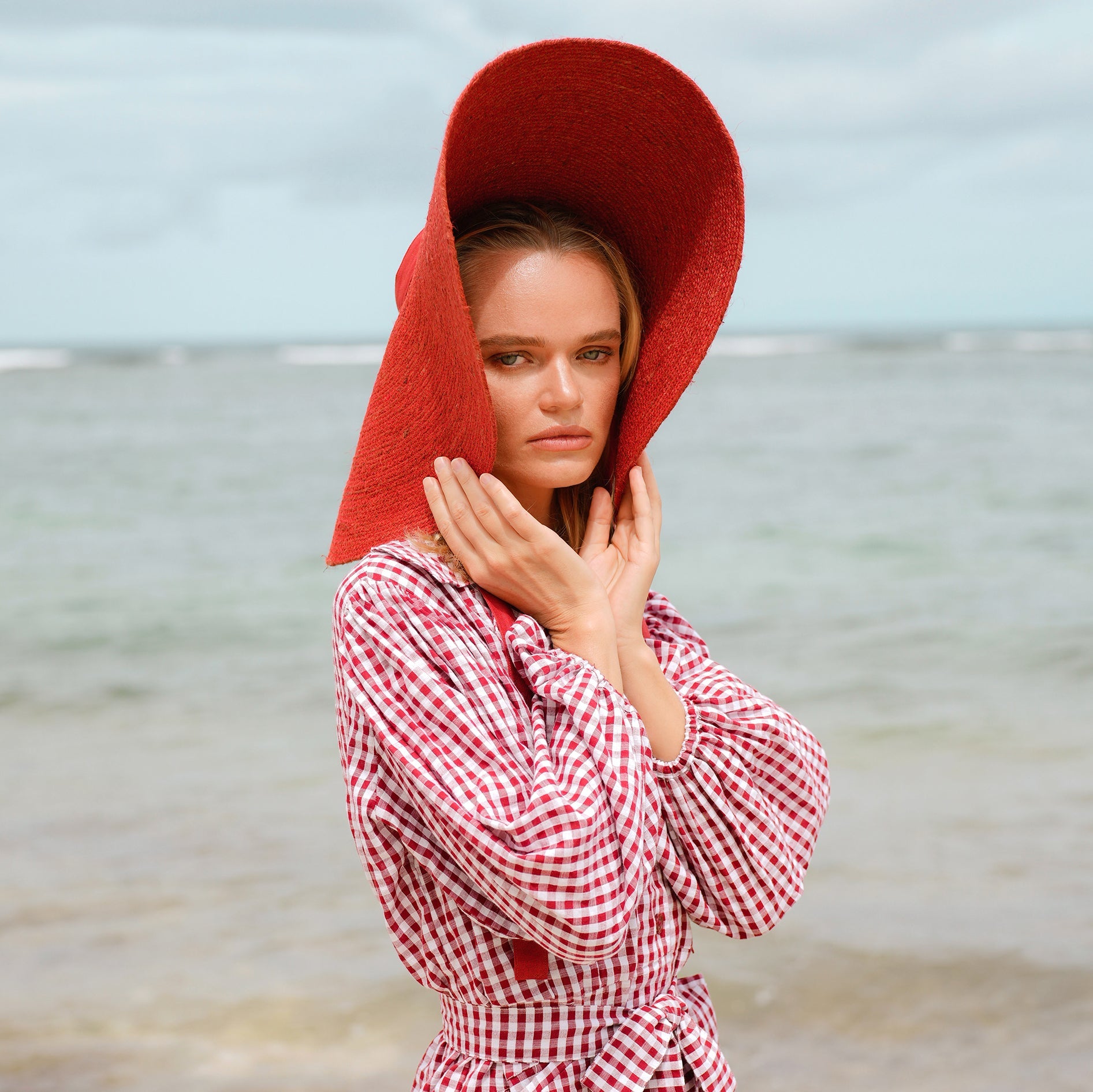 Woman wearing a red hat and checkered dress standing on a beach.