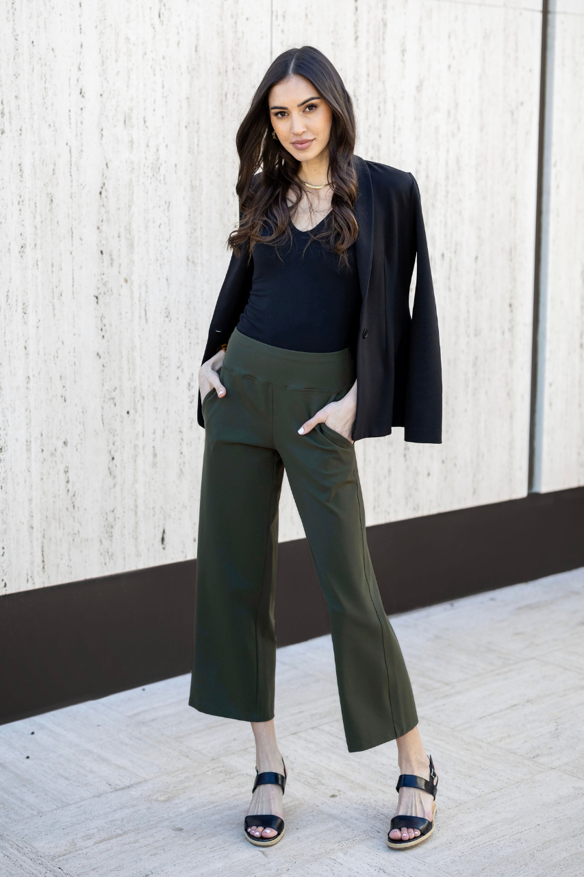 Woman wearing a black blazer and green pants standing against a light wooden wall.