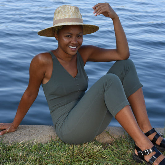 Woman sitting by water wearing a green jumpsuit and straw hat