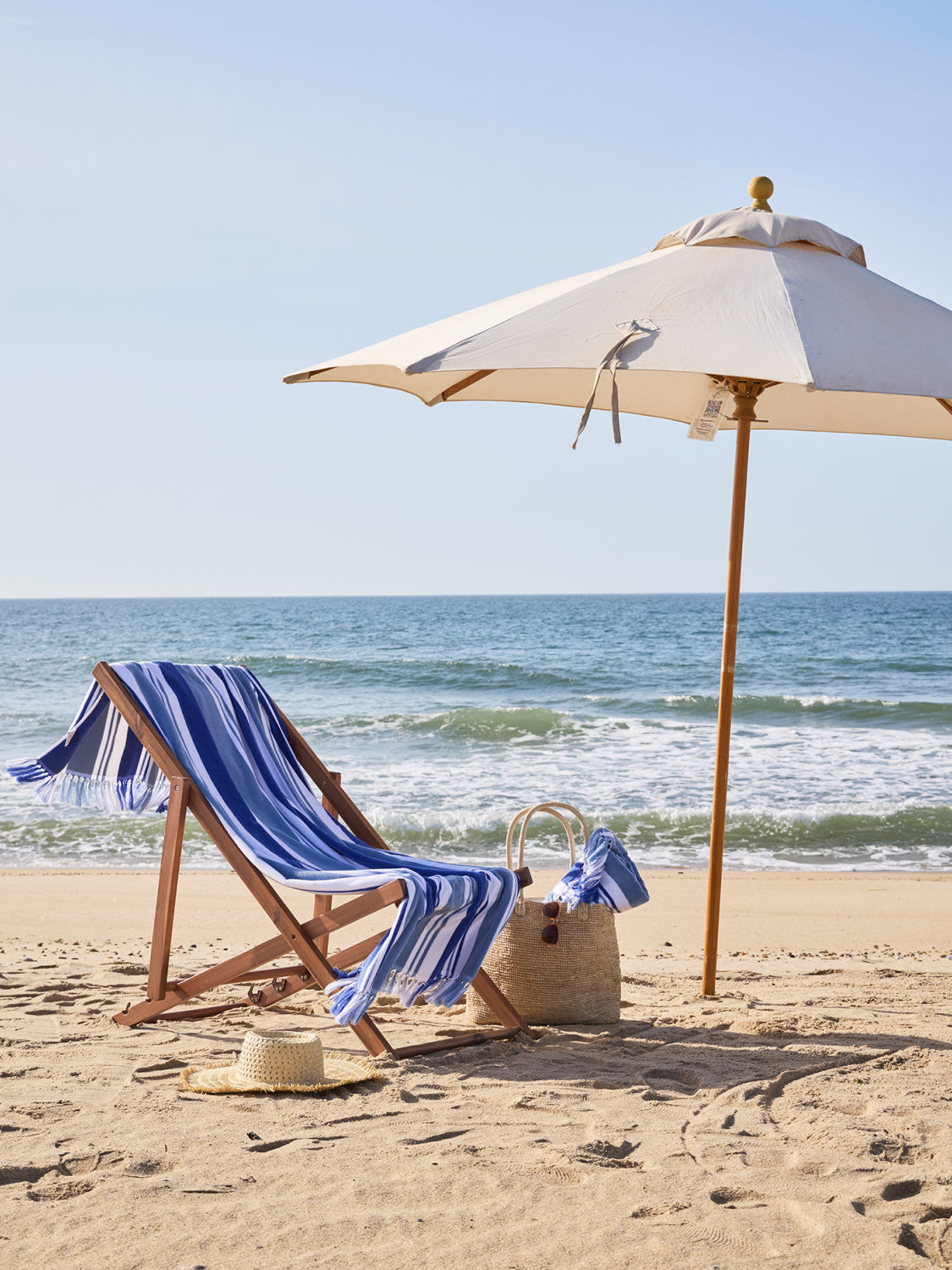 Beach chair with striped towel, umbrella, and basket on a sandy beach with ocean view.