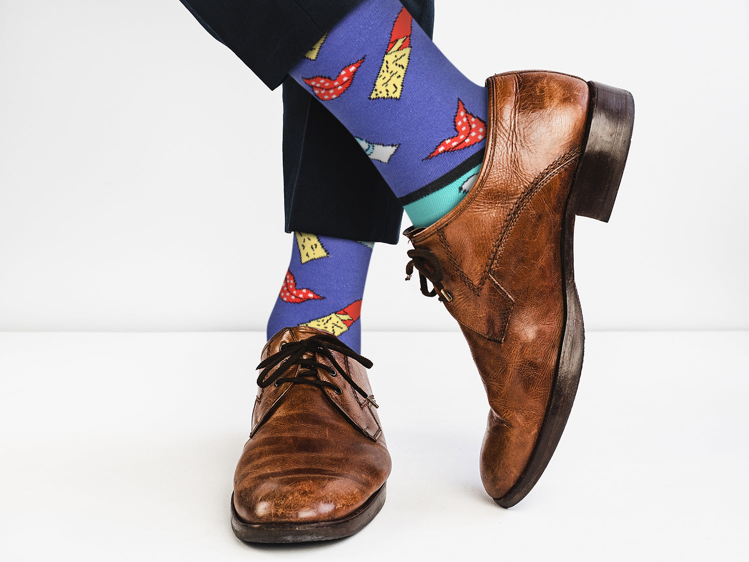 Brown dress shoes with colorful socks featuring a pattern of red and yellow shapes on a white background.