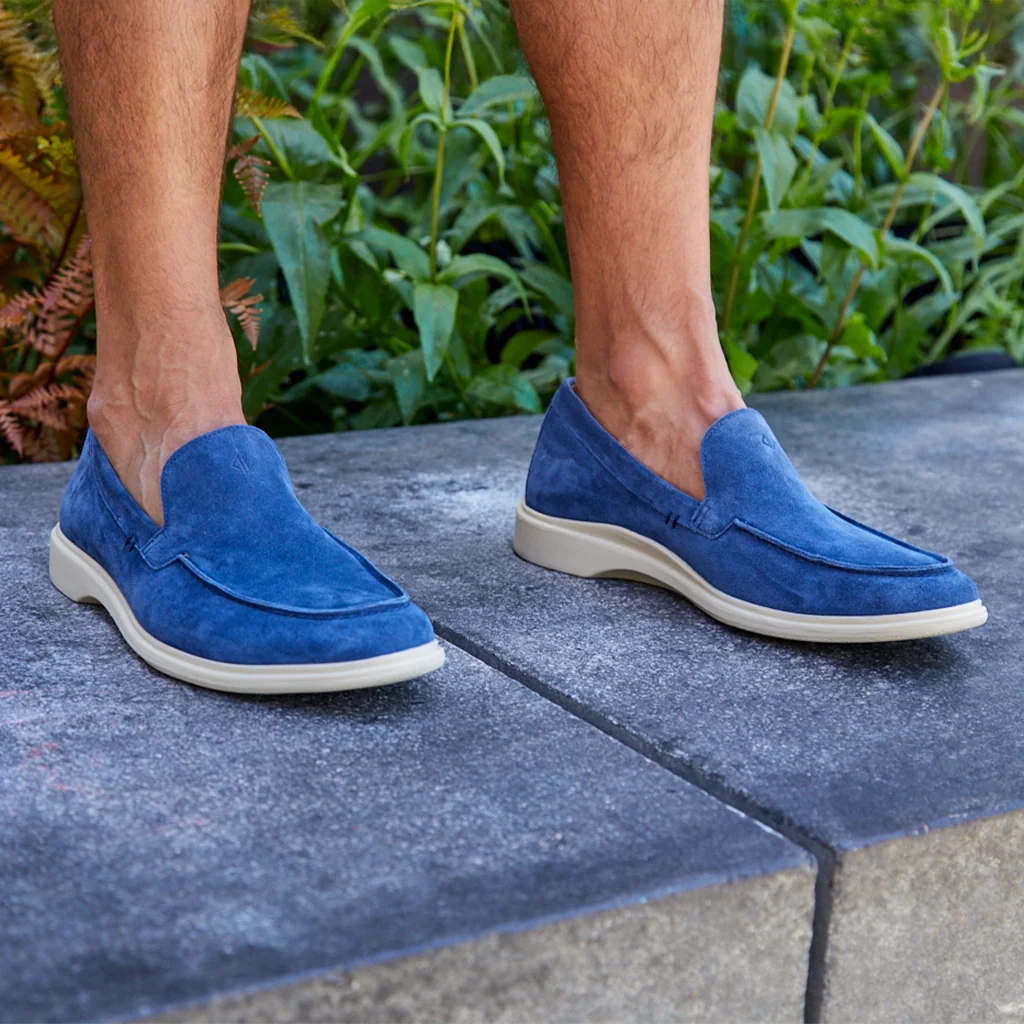 Blue loafers worn by a person standing on a stone surface with greenery in the background