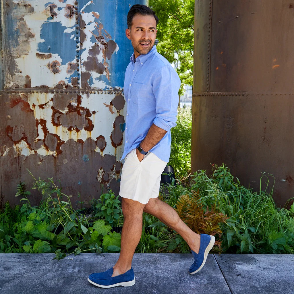 Man in a blue shirt and white shorts walking outdoors with a rustic wall and greenery in the background