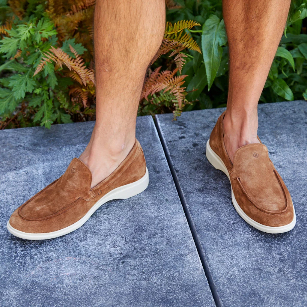 Brown suede loafers worn outdoors on a concrete surface with greenery in the background