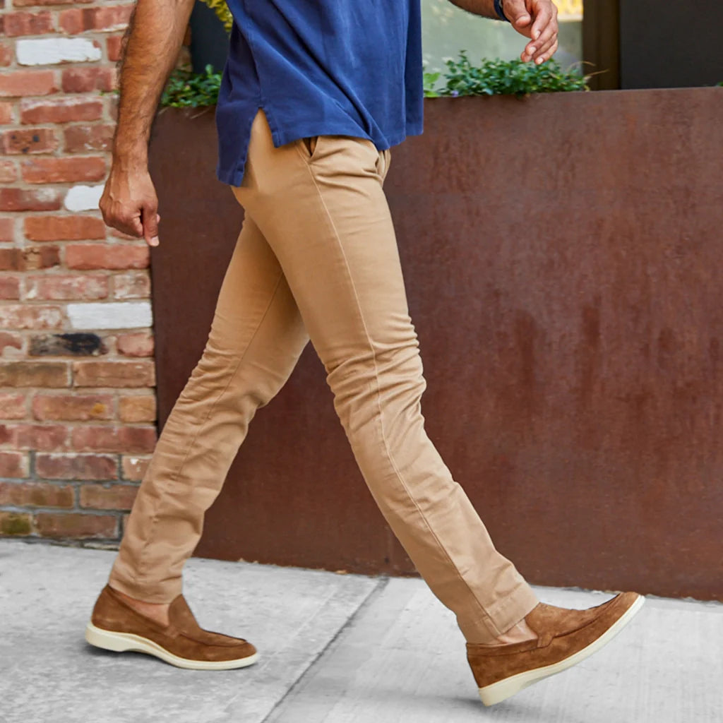 Person wearing beige pants and brown shoes walking on a sidewalk with a brick wall in the background