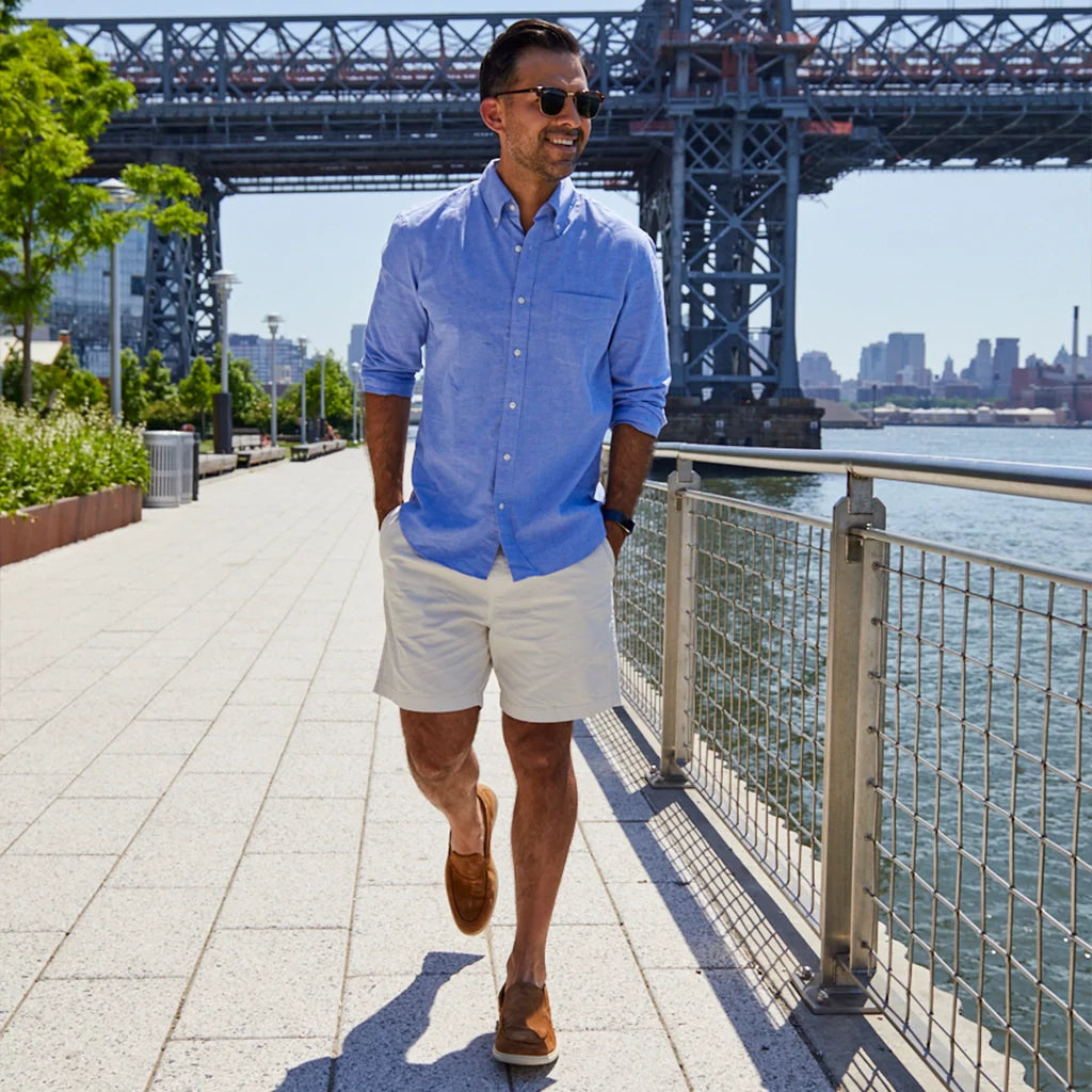 Man in a blue shirt and white shorts walking along a waterfront promenade with a bridge in the background.