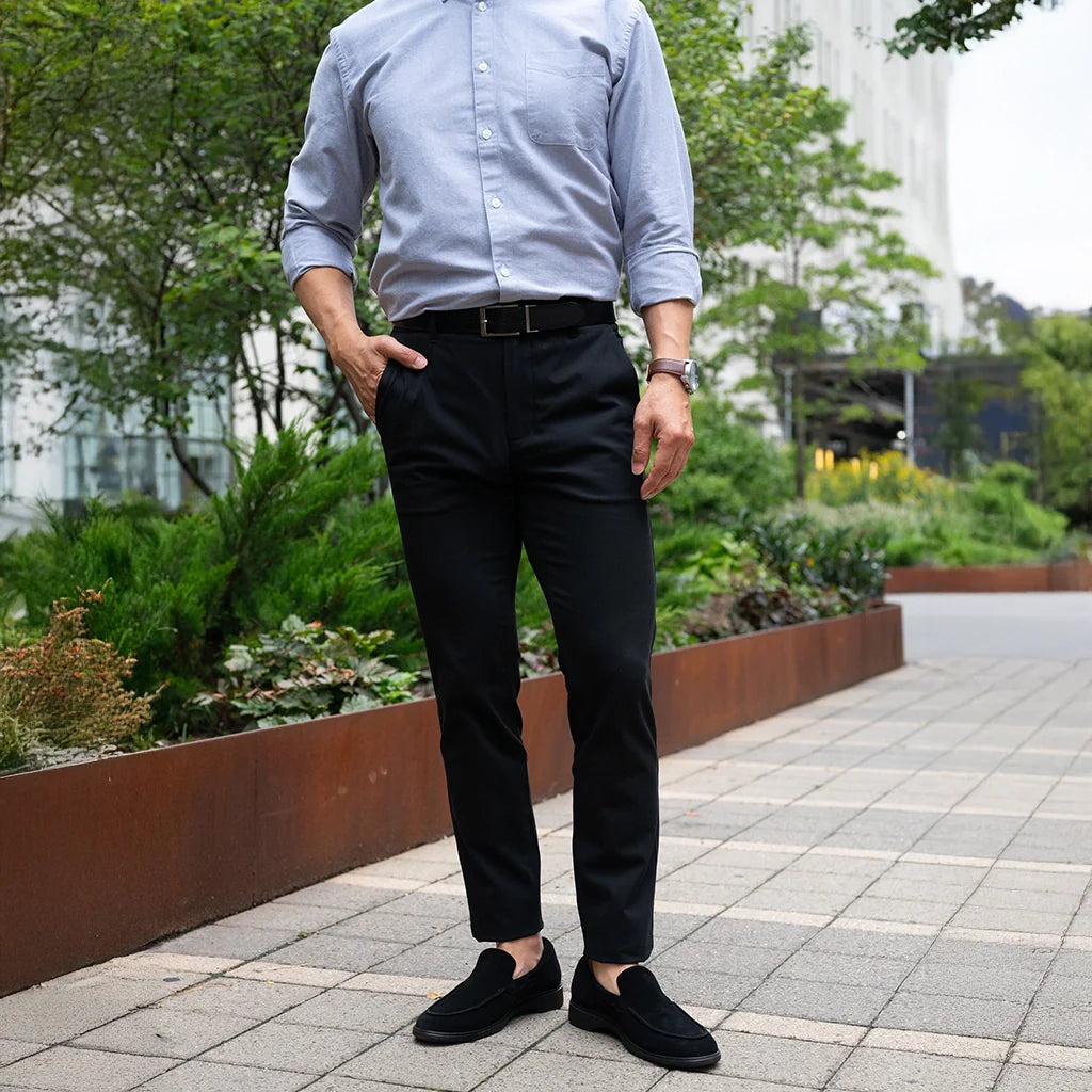 Person wearing a light blue shirt, black pants, and black shoes on a sidewalk with greenery in the background.
