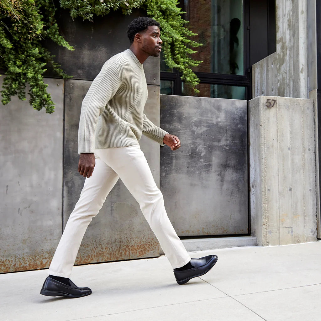 Man walking outdoors wearing a light sweater, white pants, and black shoes.