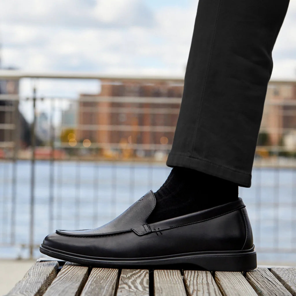Black loafer shoe worn by a person standing on a wooden platform with a blurred waterfront background.