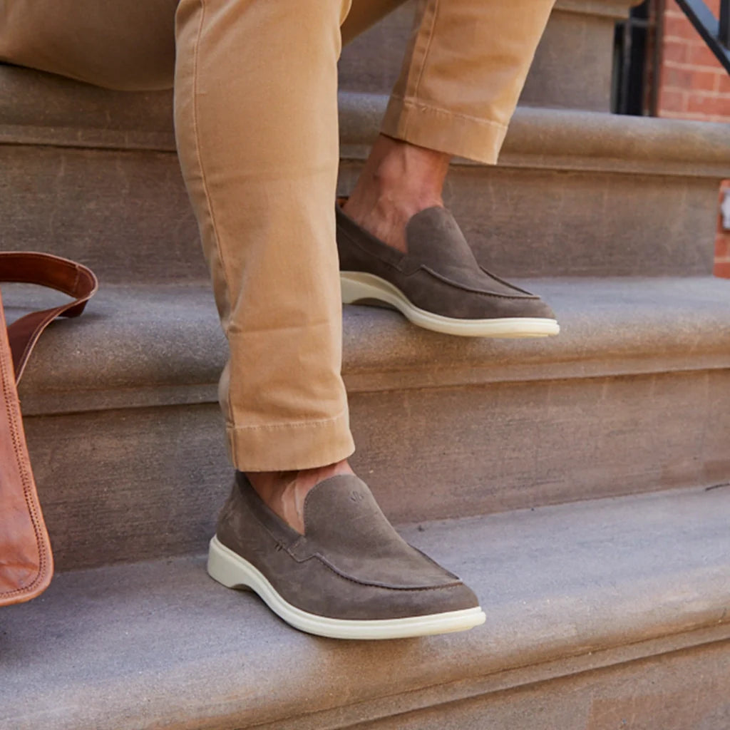 Person wearing brown loafers on stone steps with a tan bag beside them