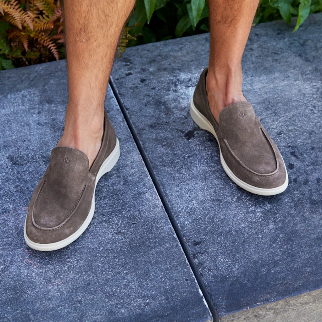 Person wearing brown loafers on a concrete surface with greenery in the background