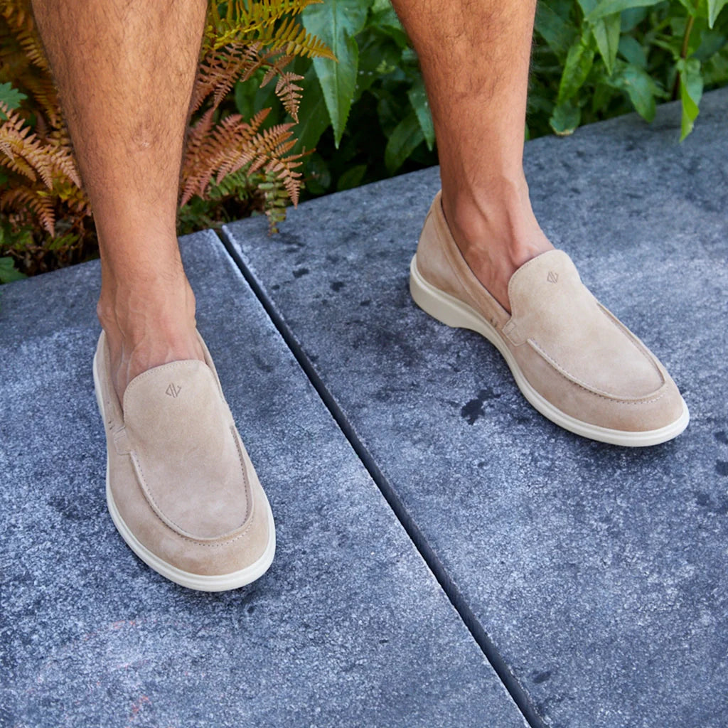 Beige loafers worn on a person's feet standing on a concrete surface with greenery in the background.