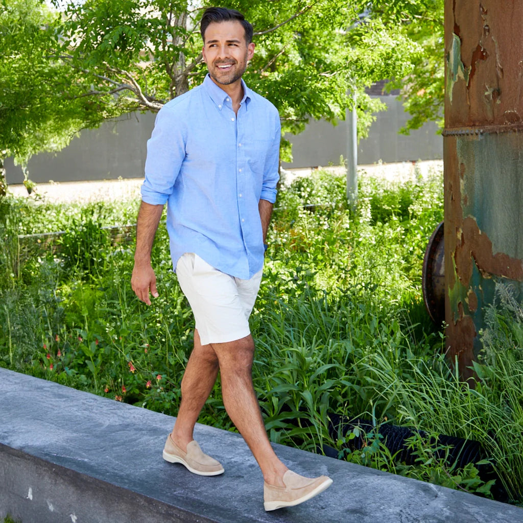 Man in light blue shirt and white shorts walking outdoors with greenery in the background