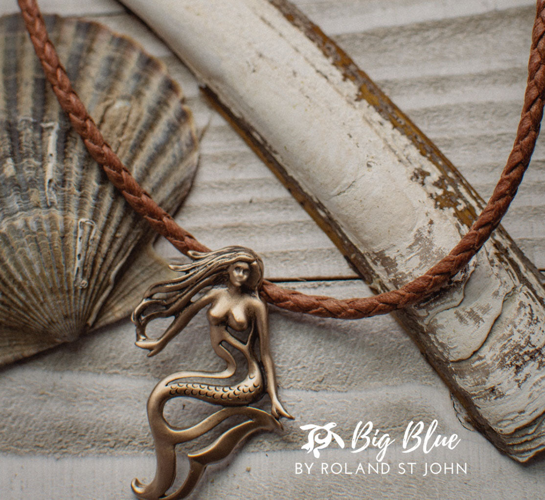 Bronze mermaid pendant on a brown cord with a shell and stone background, branded 'Big Blue by Roland St. John'.