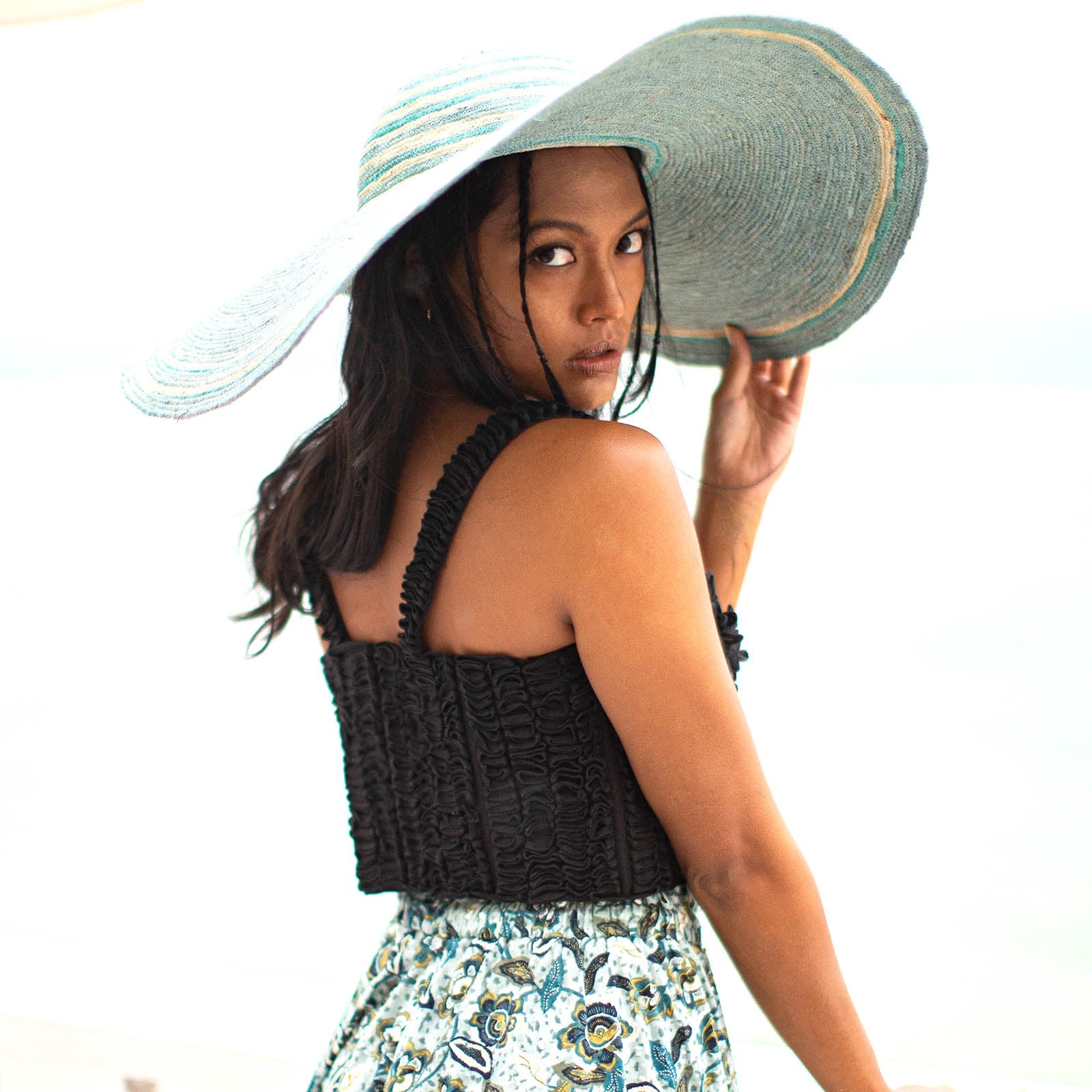 Woman wearing a black crochet top and floral skirt with a large sun hat on a white background