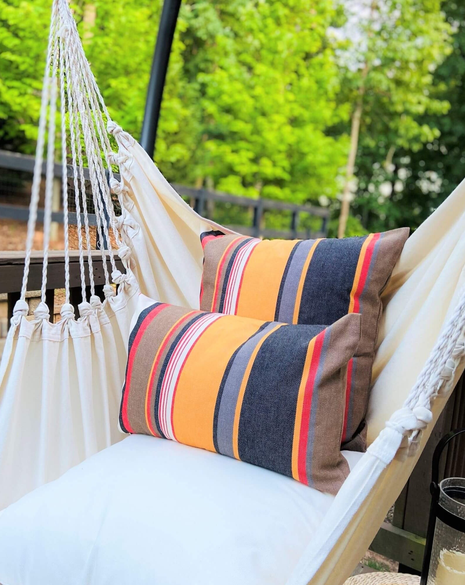 Hammock with striped pillows and a cup holder on a wooden deck with greenery in the background