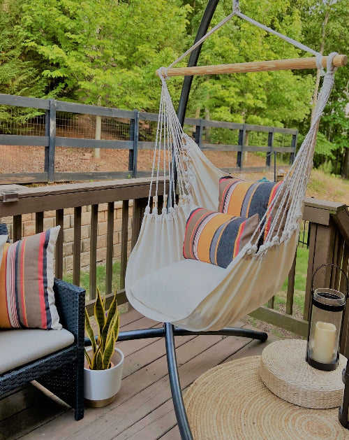 White hammock chair with colorful pillows on a wooden deck with greenery in the background