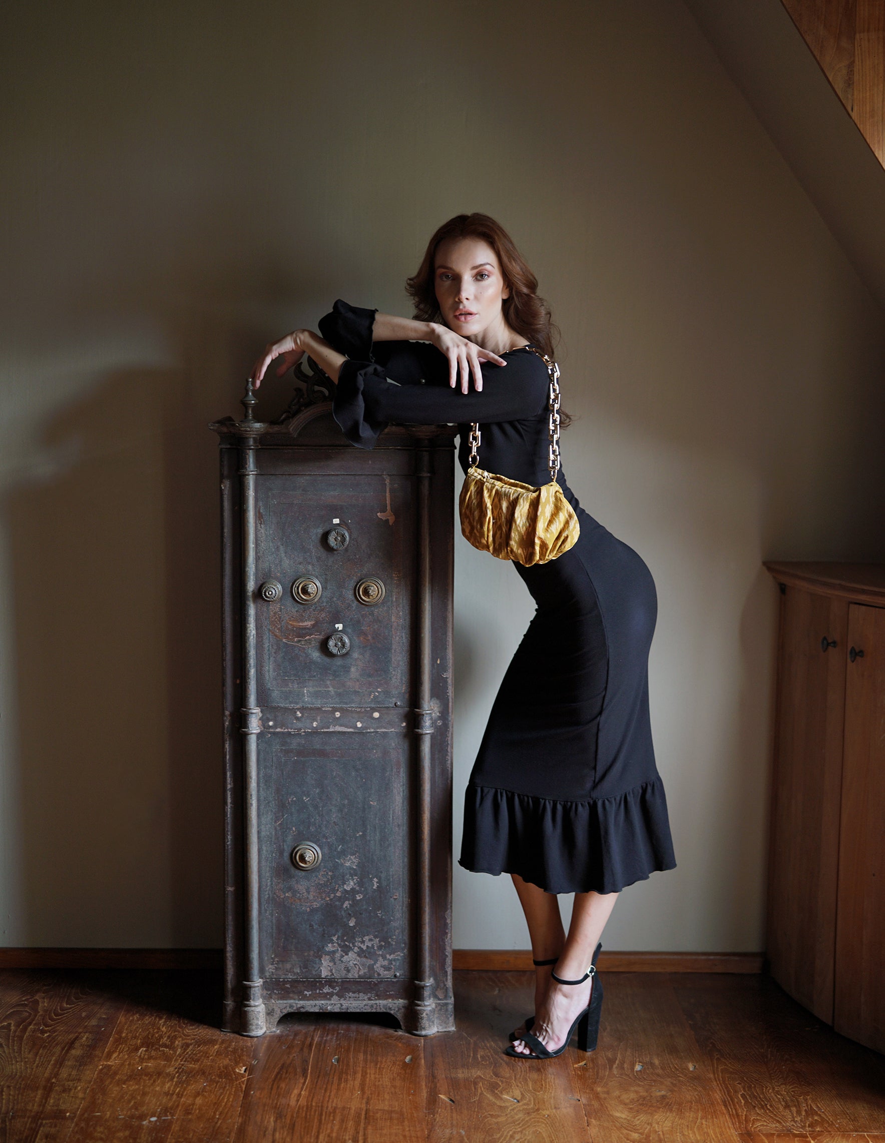 Woman in a black dress standing next to an old wooden cabinet in a dimly lit room.