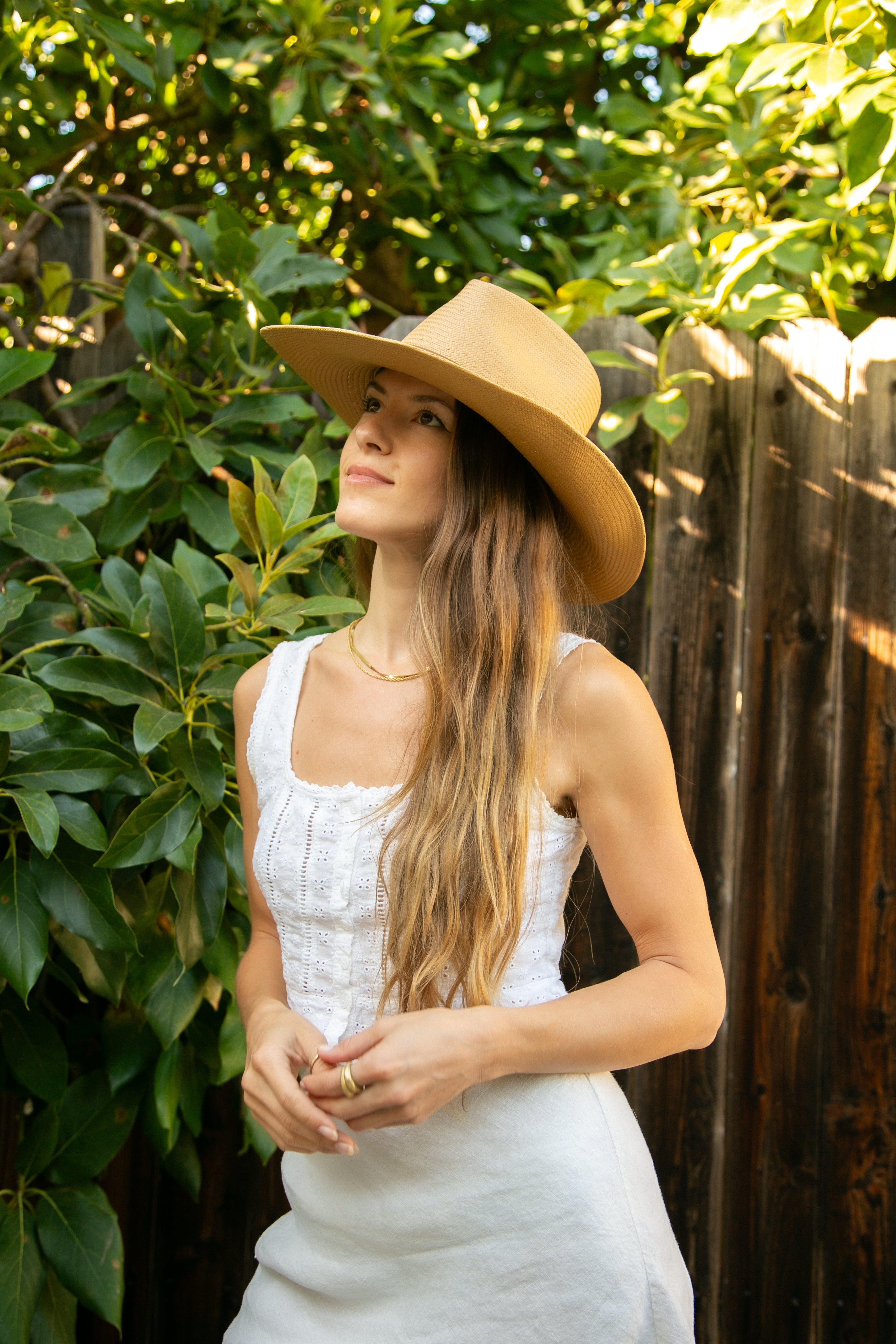 Woman wearing a wide-brimmed hat and white dress standing outdoors with greenery in the background