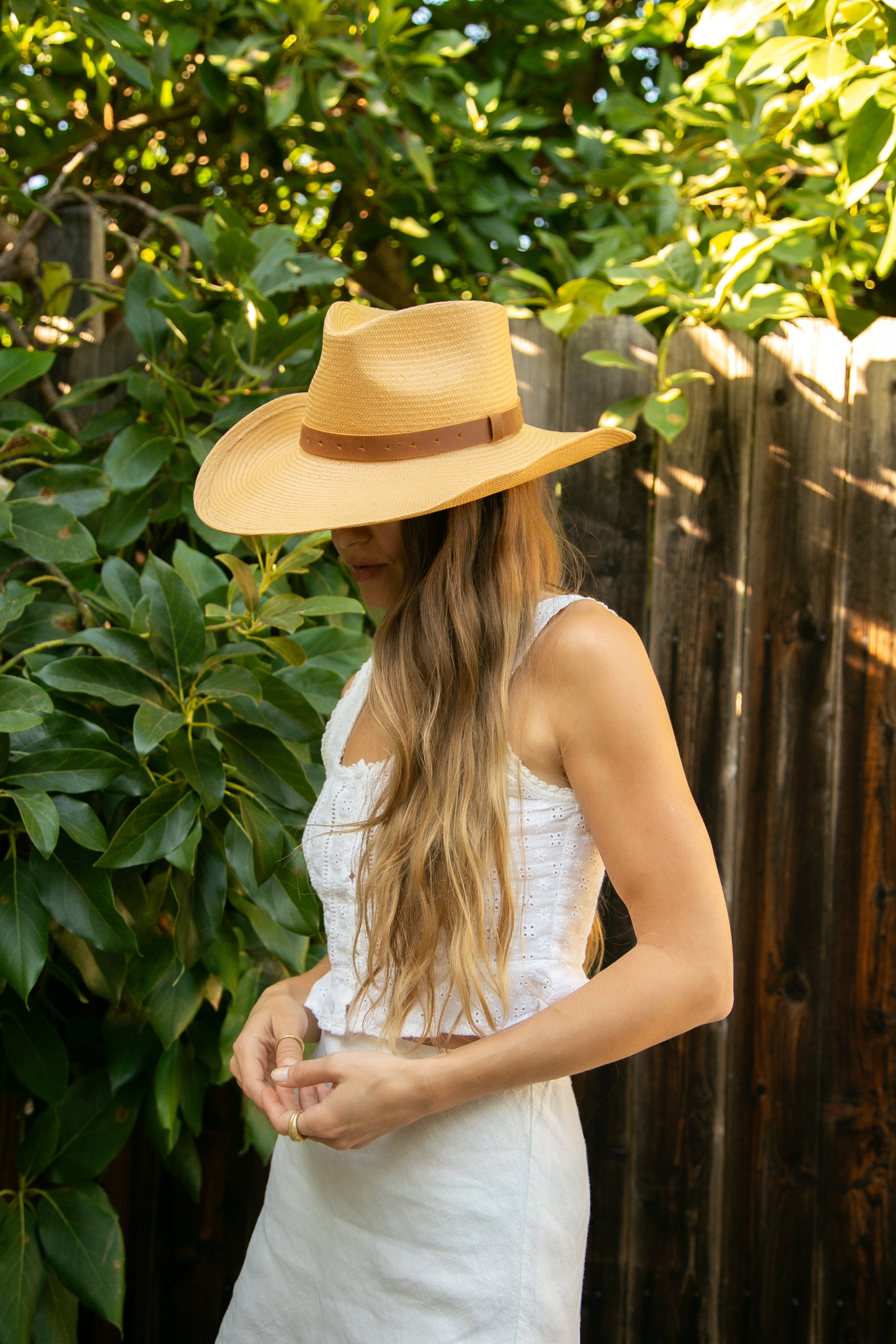 Woman wearing a straw hat and white dress standing outdoors with greenery and a wooden fence in the background.