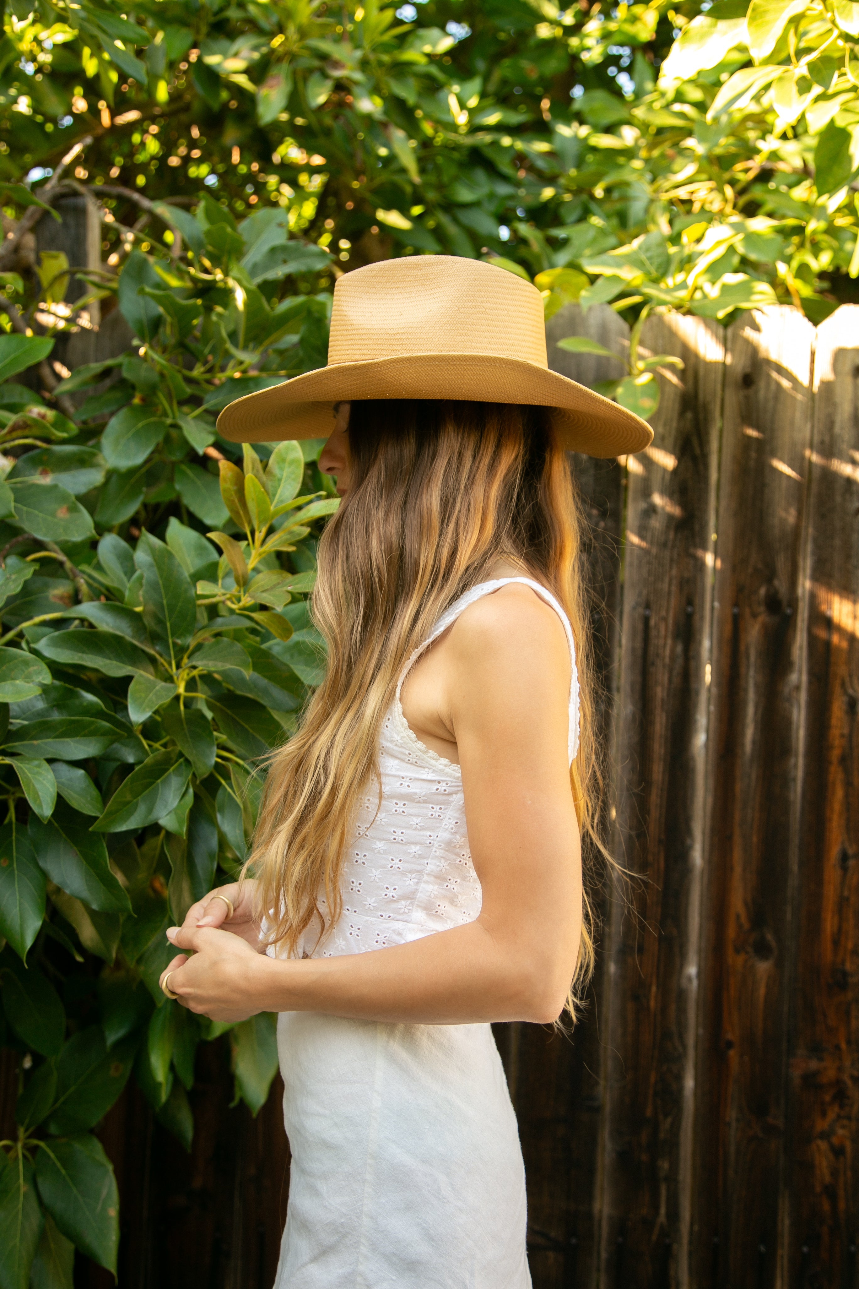 Woman wearing a beige hat and white dress standing against a wooden fence with greenery.
